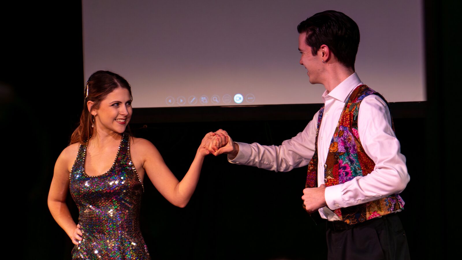 Two performers share a playful hand-hold moment on stage during Votre Toast