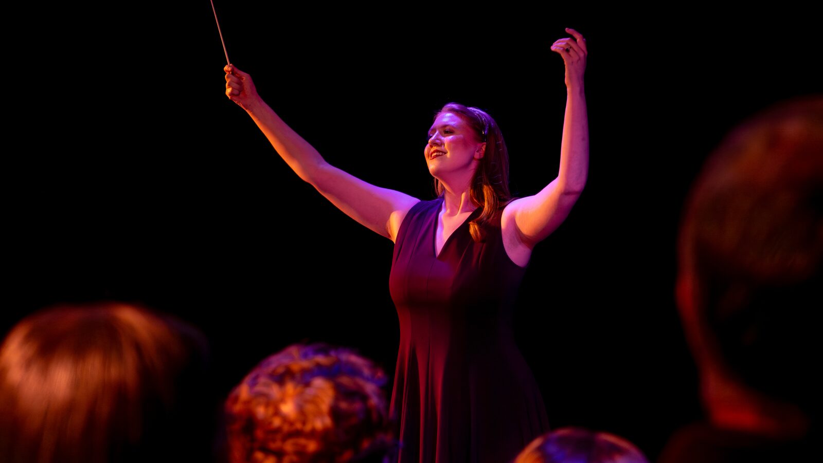 A conductor raises her baton mid-performance, audience members silhouetted in the foreground